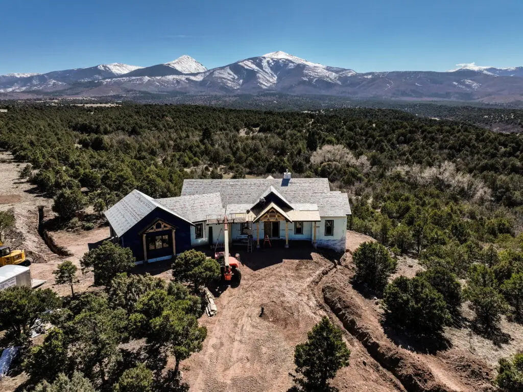 An aerial, high-angle shot shows a large custom home under construction on a dirt lot, surrounded by a dense evergreen forest with snow-capped mountains in the distance under a clear blue sky. The house features a complex roofline with grey shingles, light-colored siding, and dark blue accents on the gabled ends. A red cherry picker and other construction equipment are parked in the dirt clearing in front of the house, where trenches have been dug into the earth. The surrounding landscape is filled with rolling hills of green trees, leading up to a prominent, snow-covered mountain range on the horizon.