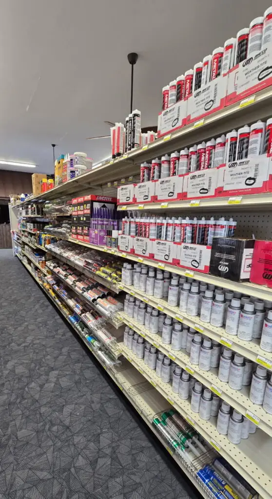 Gemini said A long aisle in a hardware store is filled with rows of neatly organized shelves. The shelves are stocked with various construction adhesives, sealants, and spray cans, including brands like Loctite and Metal Pro. The products are primarily packaged in tubes and canisters, arranged by type and color on white metal shelving over a grey patterned carpet.