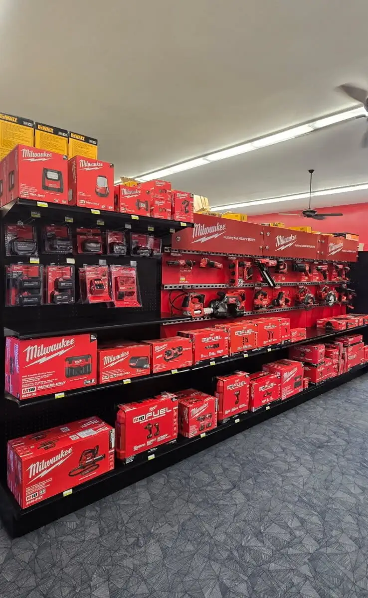 A hardware store display filled with Milwaukee power tools and accessories, featuring red boxed tool kits on shelves and hanging packaged items above, all under bright fluorescent lighting with a patterned floor beneath