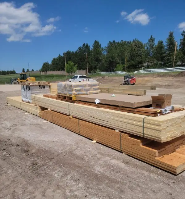 A large stack of lumber and building supplies sits on a dirt construction site under a bright blue sky with scattered clouds. The supplies include several long bundles of wood framing studs and larger beams, along with pallets of bagged concrete or mortar and boxes of hardware. In the background, a white pickup truck, a yellow front loader, and a red skid steer are visible near a line of evergreen trees and a white fence.
