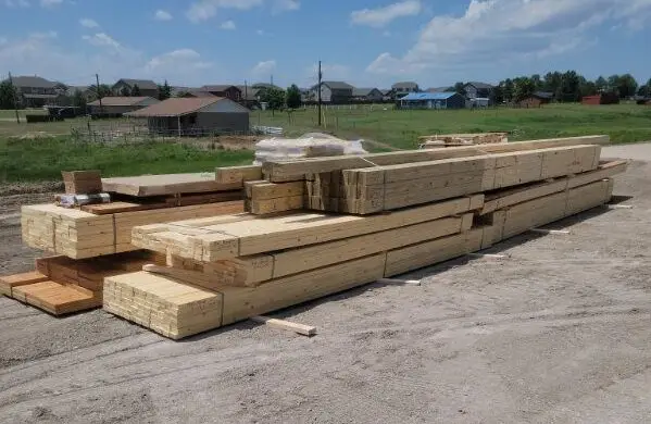 A large pile of stacked lumber and wood beams sits on a gravel lot at a construction site. The wood varies in size, including long framing studs and thicker structural beams, neatly organized in bundles. In the background, a residential neighborhood with several houses and a green field is visible under a blue sky with soft, white clouds.