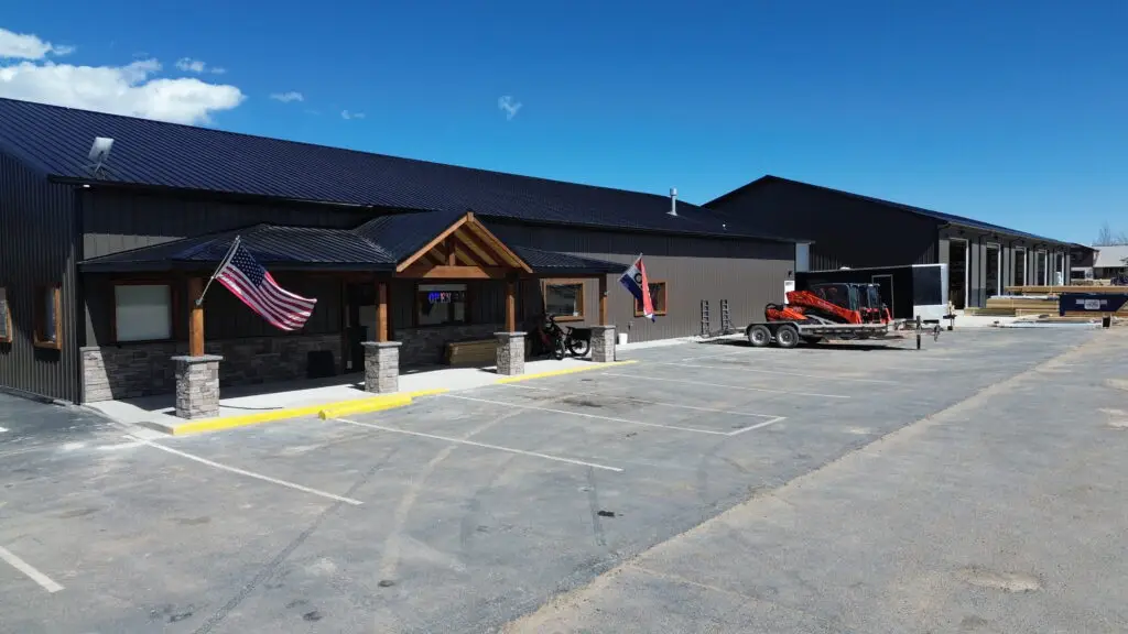 A modern industrial building with dark metal siding and a wooden gabled entrance supported by stone columns, flanked by two American flags. An empty paved parking lot sits in front, and a trailer carrying construction equipment is parked to the right under a clear blue sky.