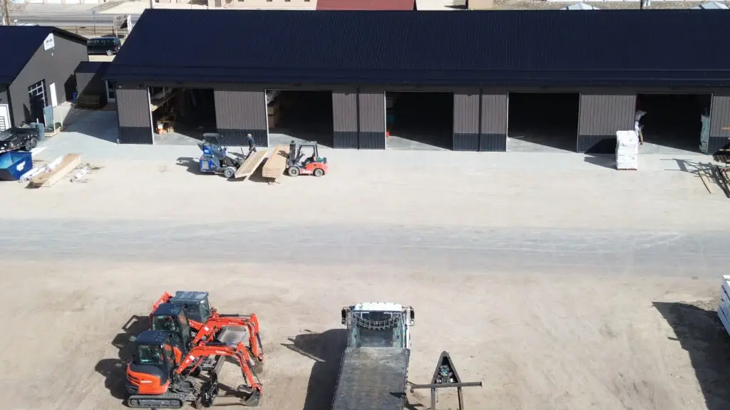 An aerial view shows a lumber yard and warehouse facility with a large, dark-grey building featuring multiple open bay doors. In the foreground, heavy machinery including two orange mini-excavators and a flatbed truck are parked on a dirt lot. Near the warehouse bays, two forklifts are actively moving stacks of lumber across the concrete apron.