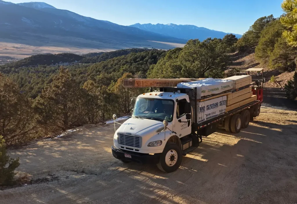 A large flatbed truck loaded with lumber and wrapped building materials parked on a dirt road, surrounded by trees and mountains under a clear blue sky.