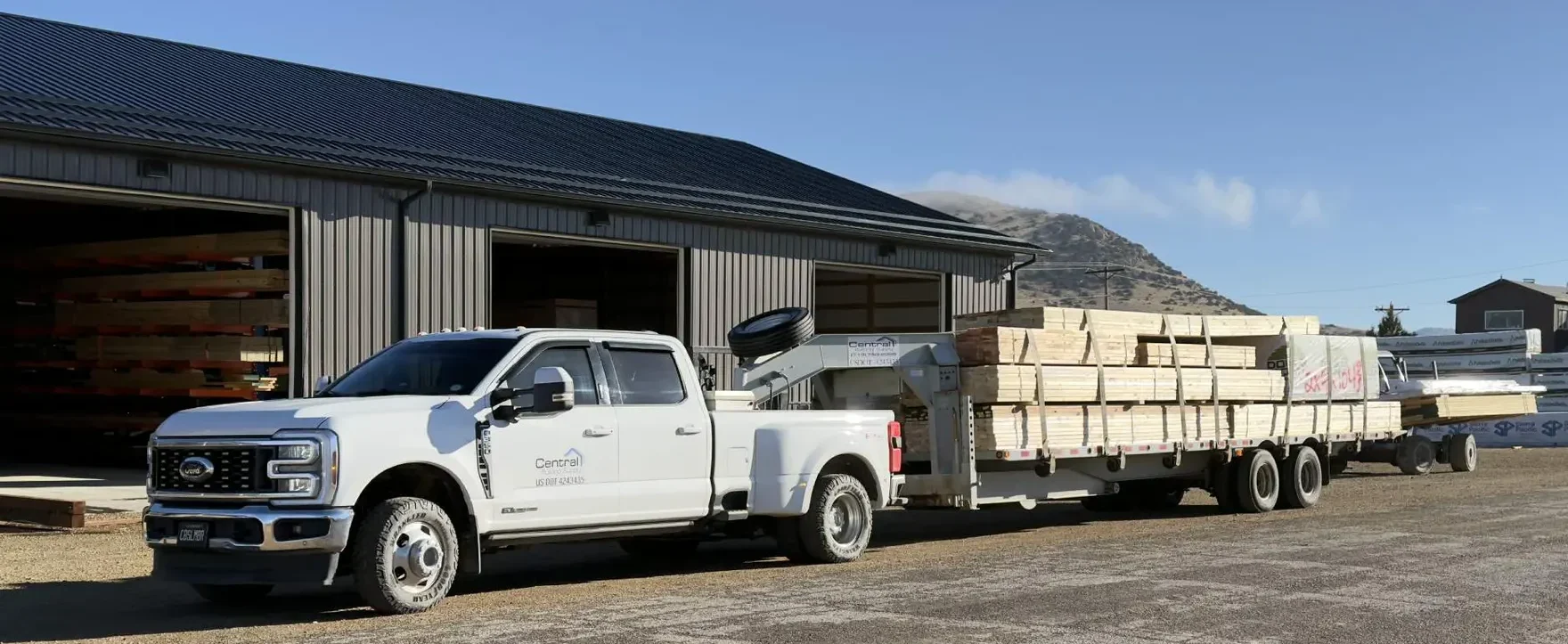 A white pickup truck hitched to a flatbed trailer loaded with stacked lumber, parked outside a metal building with open bays and additional lumber stored inside, set against a clear mountain backdrop.