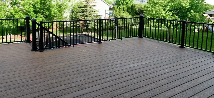 An elevated outdoor deck with composite wood flooring and black metal railings, overlooking a green yard with trees and a neighboring house in the background.