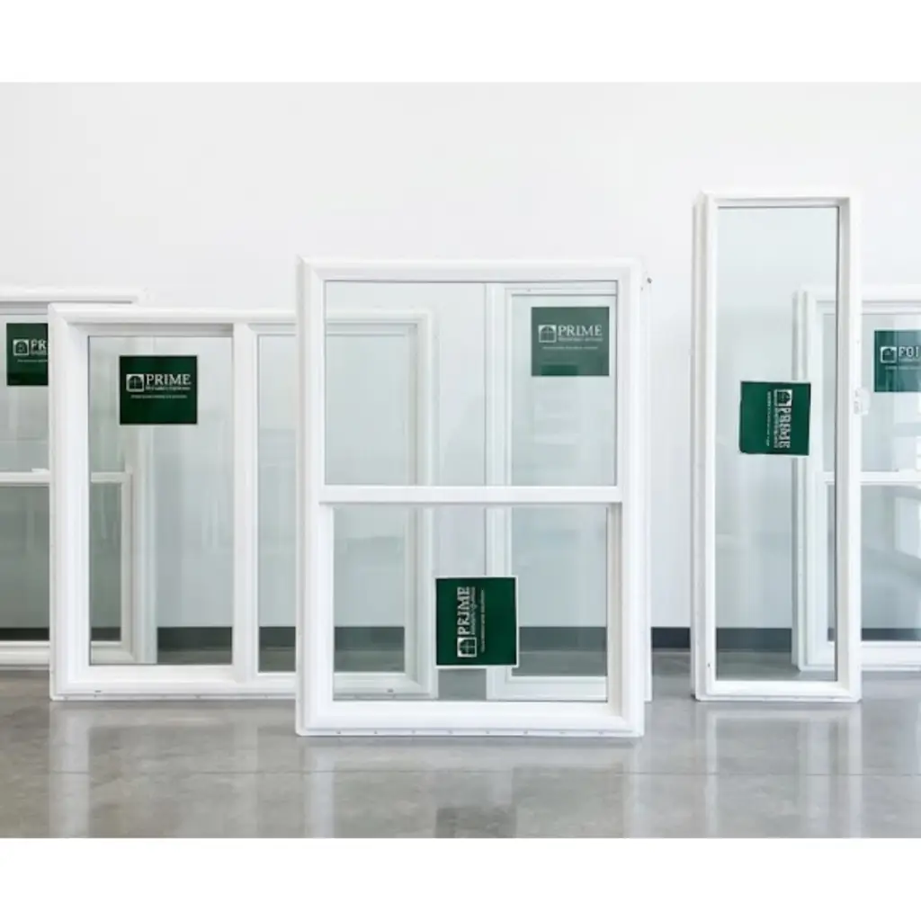 Gemini said Several white vinyl windows of different shapes and sizes are displayed on a polished concrete floor against a plain white wall. The assortment includes a narrow vertical window and wider double-hung or sliding styles. Each glass pane features a green and white Prime Window Systems sticker. The lighting is bright and even, creating a clean, professional product showcase.
