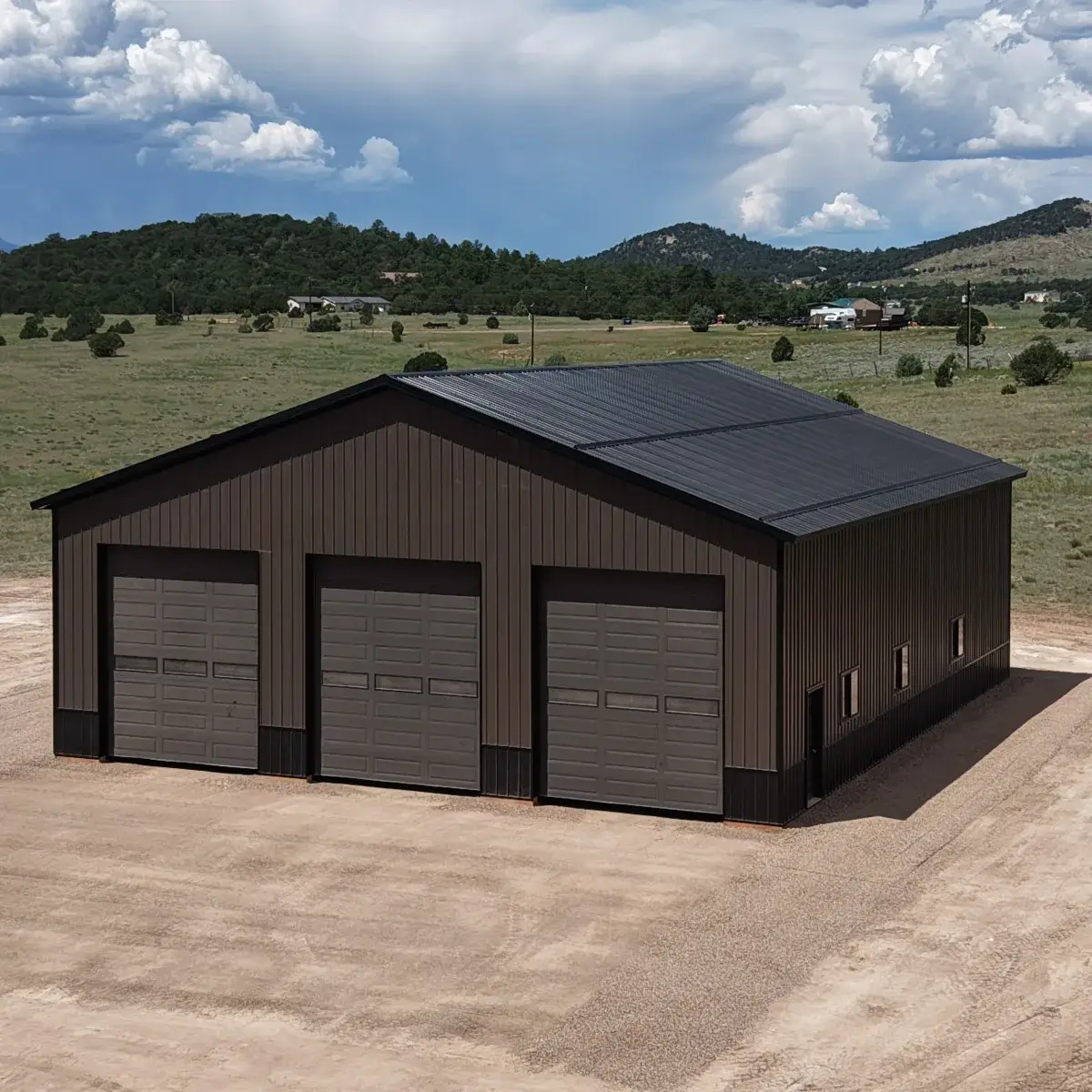 Brown metal-sided building with three overhead doors, a walk-in door and side windows in a rural landscape.